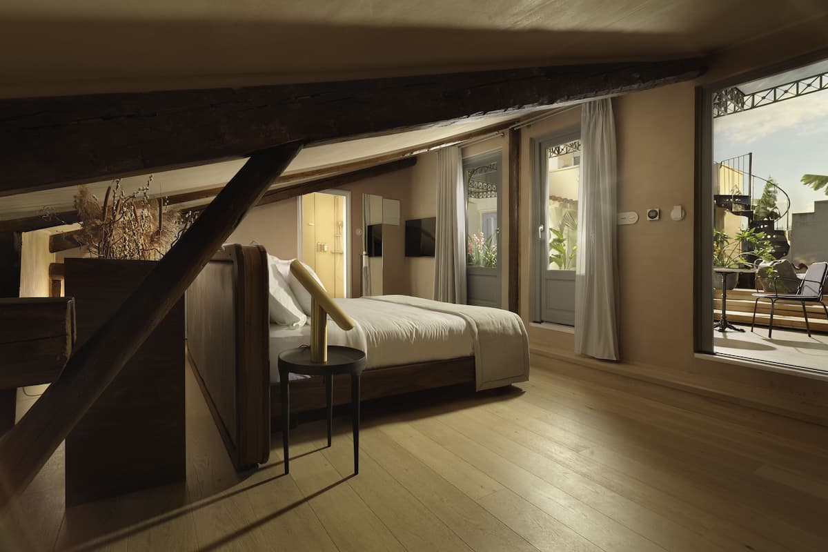Quarto com cama, uma mesinha, um vaso de planta e janela com vista para o exterior.