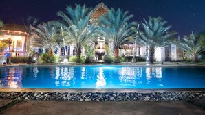 A nighttime view of a resort with a swimming pool, palm trees, and a building with a thatched roof.