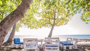 A beachside seating area with white wooden chairs and blue cushions, shaded by trees, overlooking the sea.