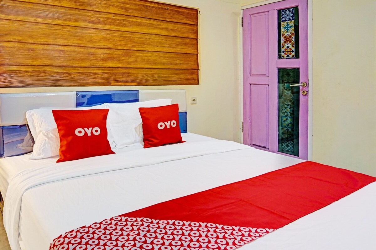A hotel room with a bed, pillows with 'OYO' branding, a wooden headboard, and a purple door with a decorative window.