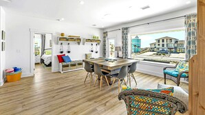 A modern living room with a wooden dining table, black chairs, a sofa with colorful cushions, a wicker chair, and a large window with a view of buildings. - Coastal Camphouse: Gulf View Home | Private Pool, Pet Friendly, Amenities+ (Navarre)