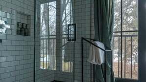 A bathroom with white tiled walls, a glass shower enclosure, a towel rack with towels, and a view of trees through a window. - Luxury Douglas Beach House (Douglas)