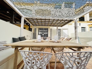 An outdoor dining area with a wooden table and white chairs, a pergola overhead, and a view of the surrounding buildings.