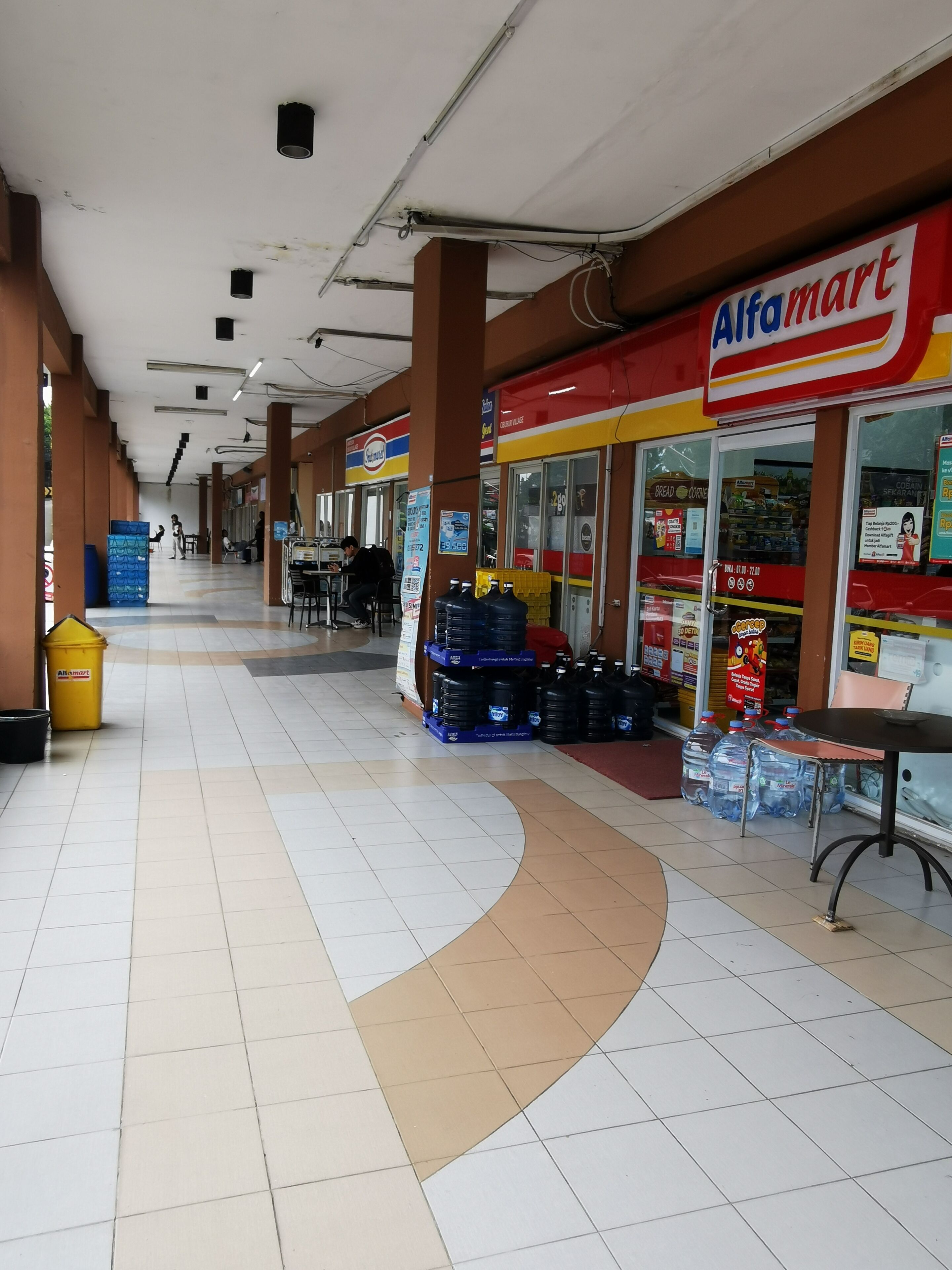 Corredor de um shopping com uma loja Alfamart à direita, piso em azulejos e teto curvo.