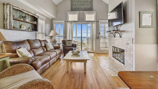 A spacious living room with a high ceiling, a fireplace, a television mounted on the wall, and a view of the outdoors through large windows. - Welcome to Stella Di Mare! (St. George Island)