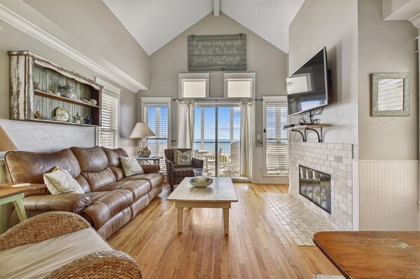 A spacious living room with a high ceiling, a fireplace, a television mounted on the wall, and a view of the outdoors through large windows.