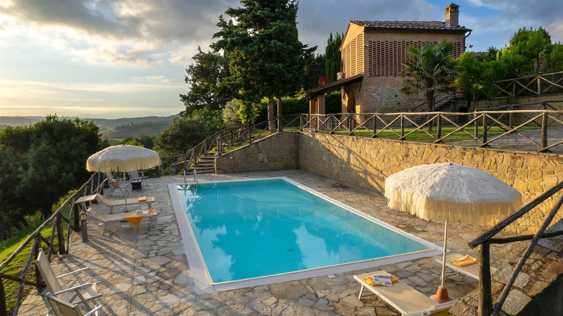 A swimming pool with stone flooring, surrounded by lounge chairs and umbrellas, with a stone wall and a building in the background.