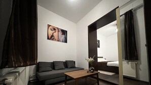 A modern, well-lit room with a dark-colored sofa, a wooden coffee table, and a painting on the wall. There is a bed with a white headboard in the background, and a window with dark curtains. - Old City Appartment (București)