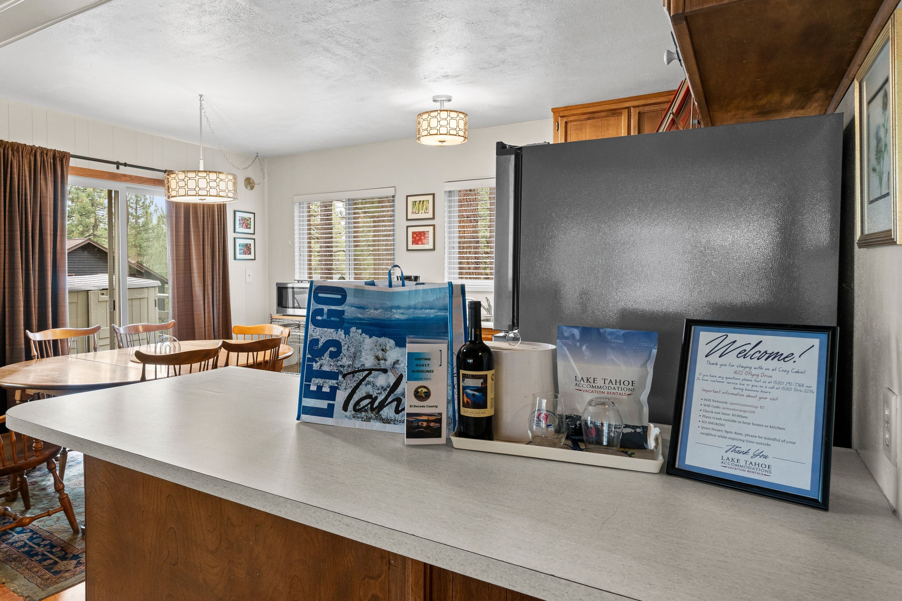 A kitchen area with a refrigerator, a dining table with chairs, a hanging light fixture, and a window with blinds.
