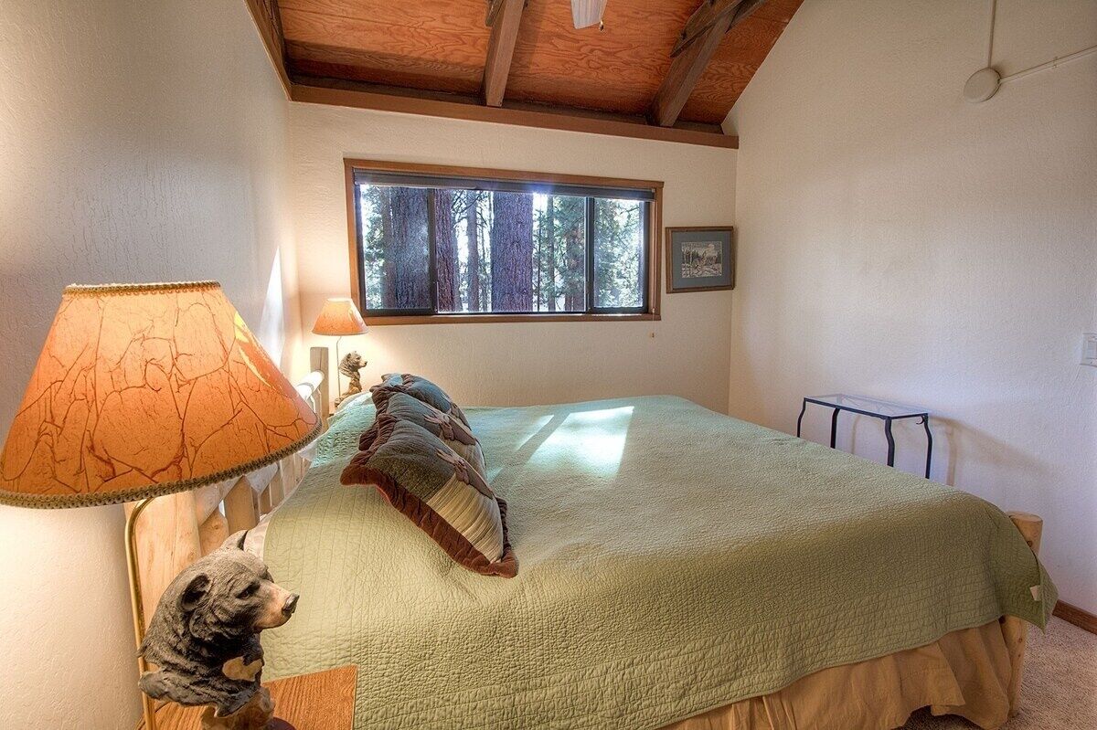 A bedroom with a bed, a lamp, a bear figurine, a window with a forest view, and a picture on the wall.