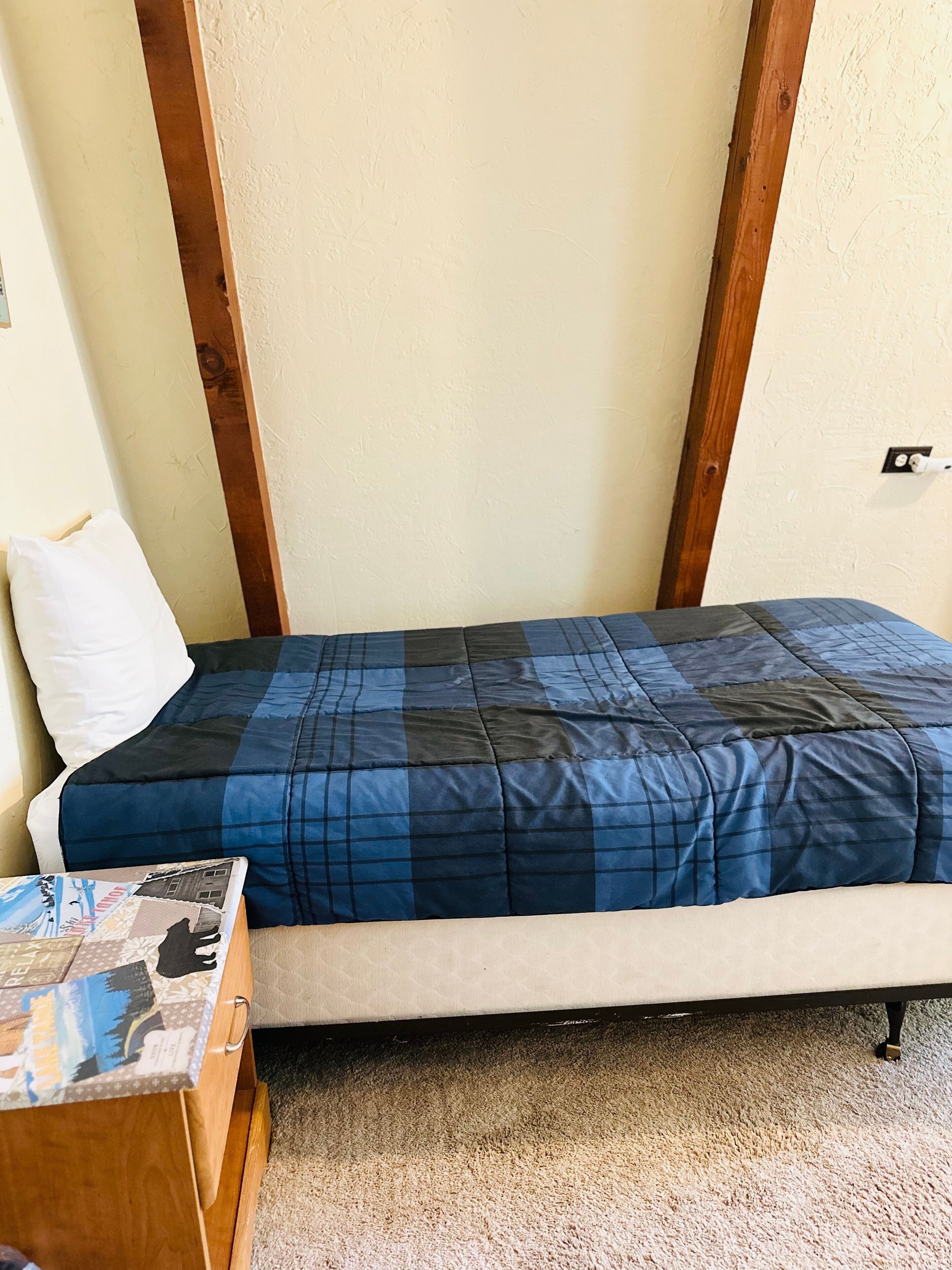 A small, single-bed room with a blue and black checkered bedspread, a white pillow, and a wooden nightstand with a magazine.