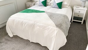 Una cama bien hecha con una colcha blanca y verde, dos mesitas de noche con lámparas y una ventana con persianas.
