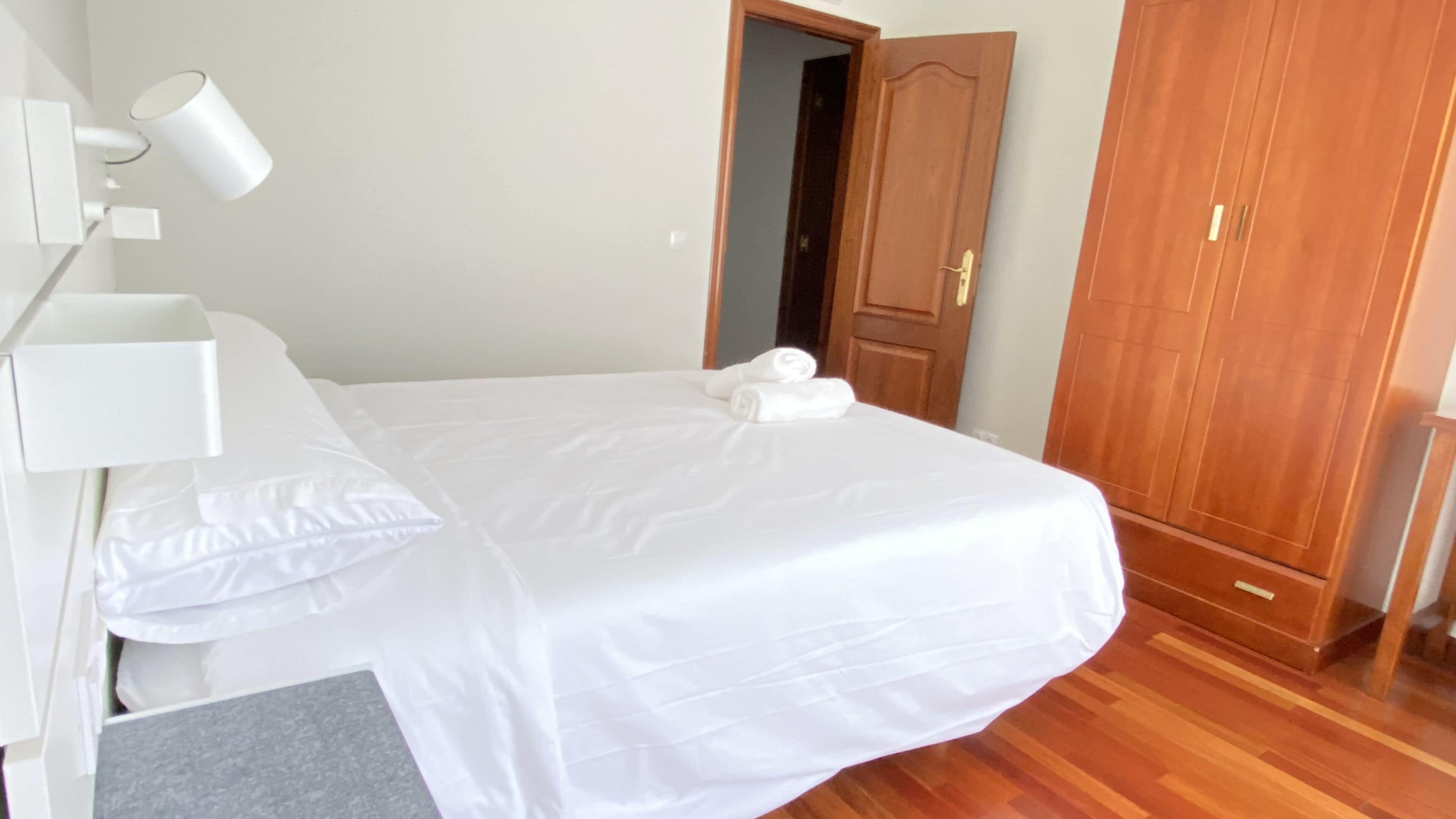 A neatly made bed with white linens and two rolled towels, a wooden wardrobe, and a white bedside table with a lamp.