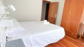 A neatly made bed with white linens and two rolled towels, a wooden wardrobe, and a white bedside table with a lamp.