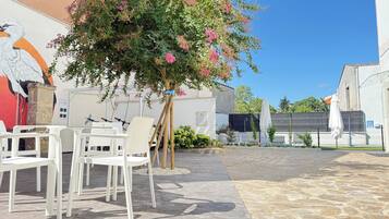 An outdoor seating area with white plastic chairs and tables, a tree with pink flowers, and a mural on the wall.