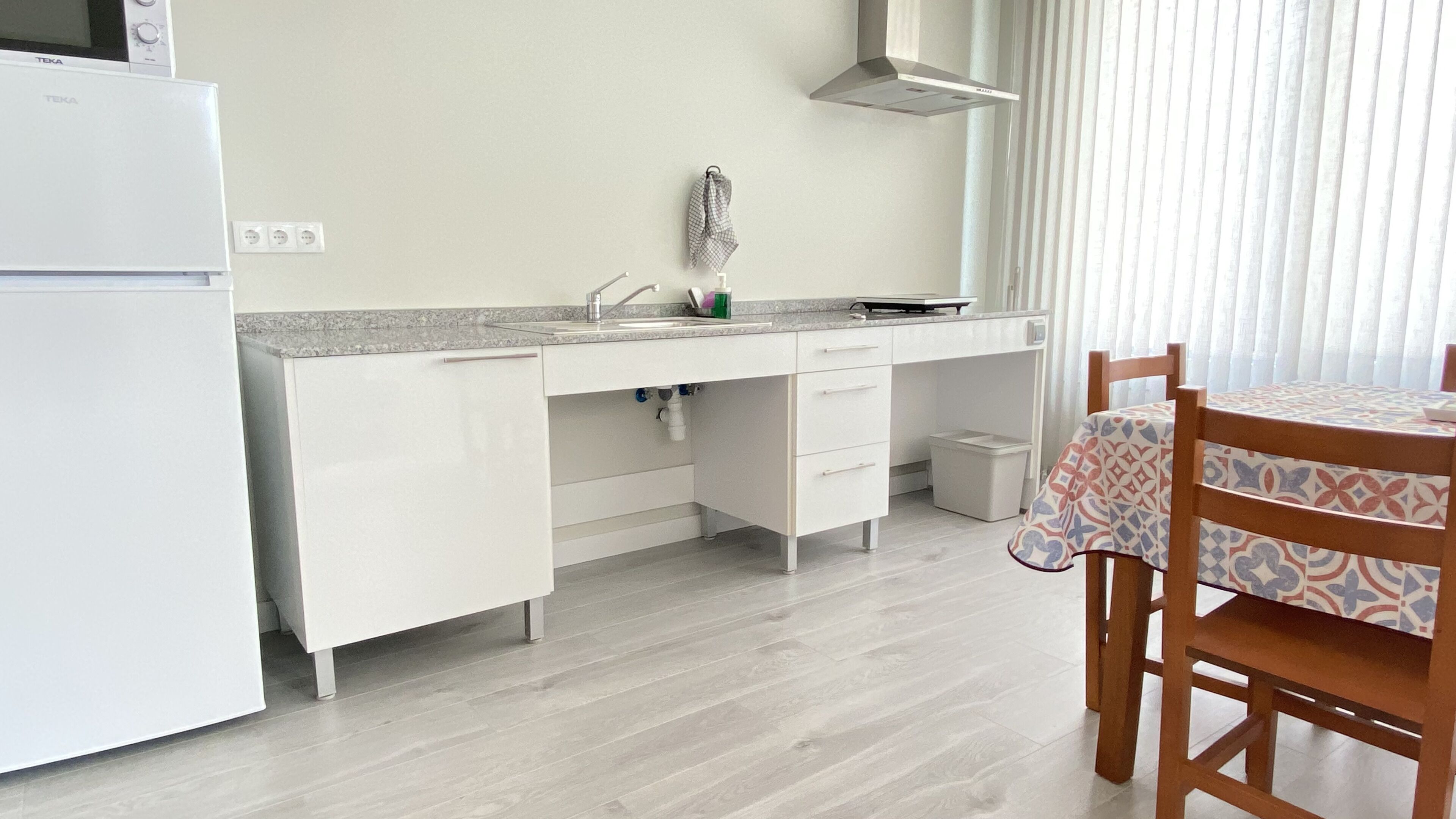 A modern kitchen with white cabinetry, a built-in microwave, and a sink. There's a small table with a patterned tablecloth and a chair.