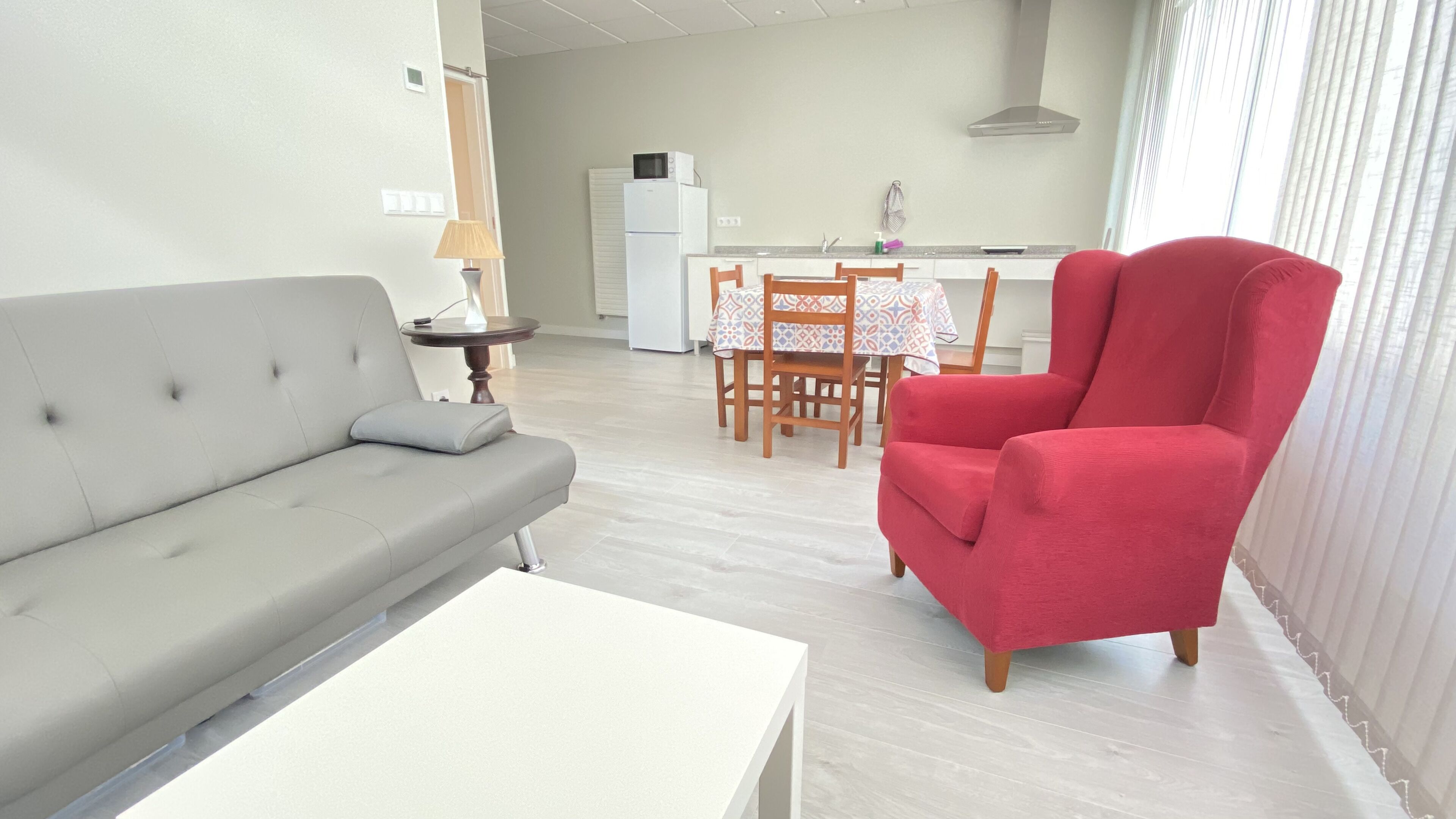 Una sala de estar moderna con un sofá gris, un sillón rojo, una mesa de centro blanca y un comedor con mesa y sillas.