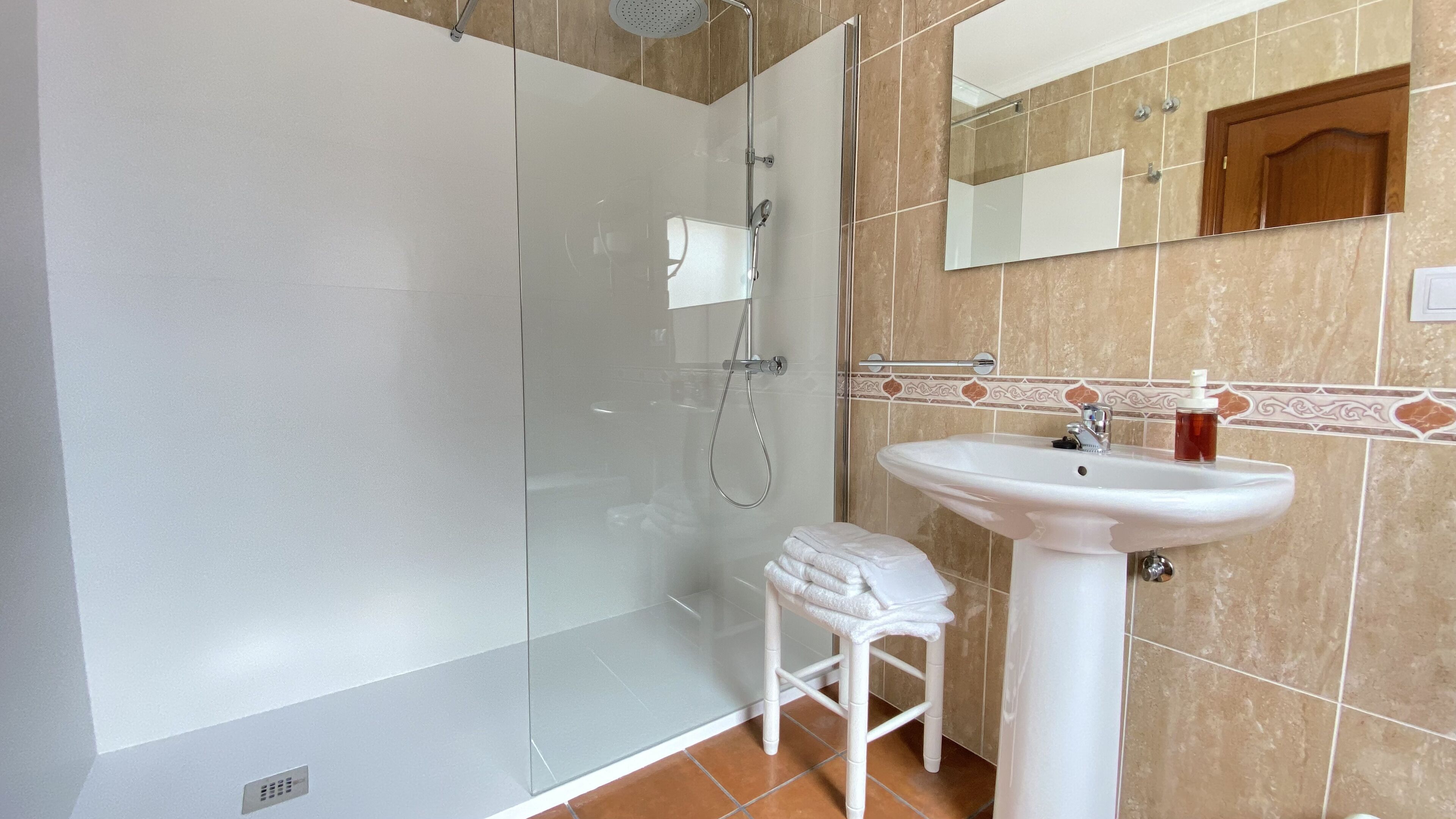 Een badkamer met een glazen douchecabine, een wit wastafelmeubel en een klein krukje.