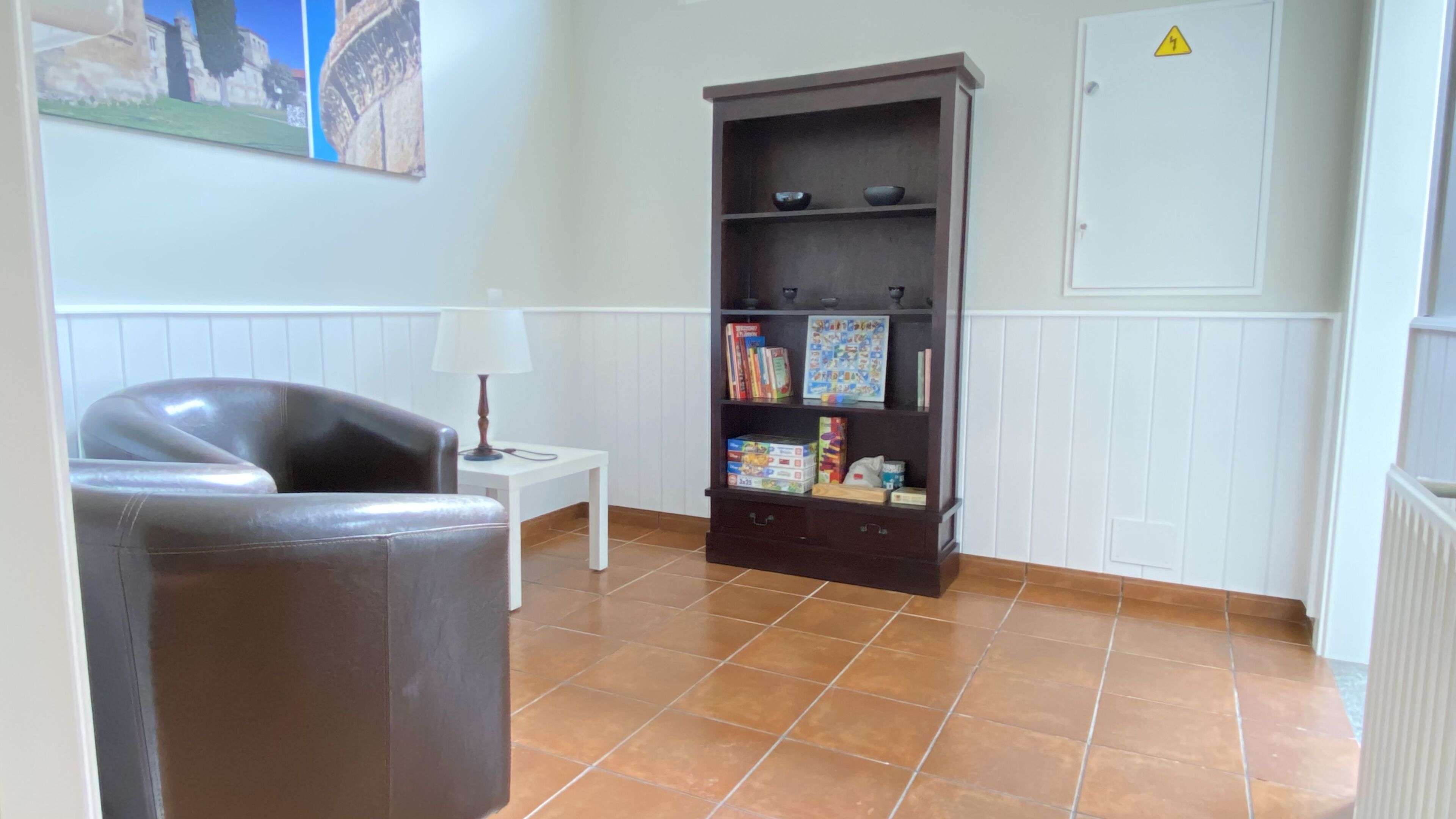 A room with a brown leather chair, a white side table with a lamp, a dark wooden bookshelf with books and games, and a white door with a yellow warning sign.