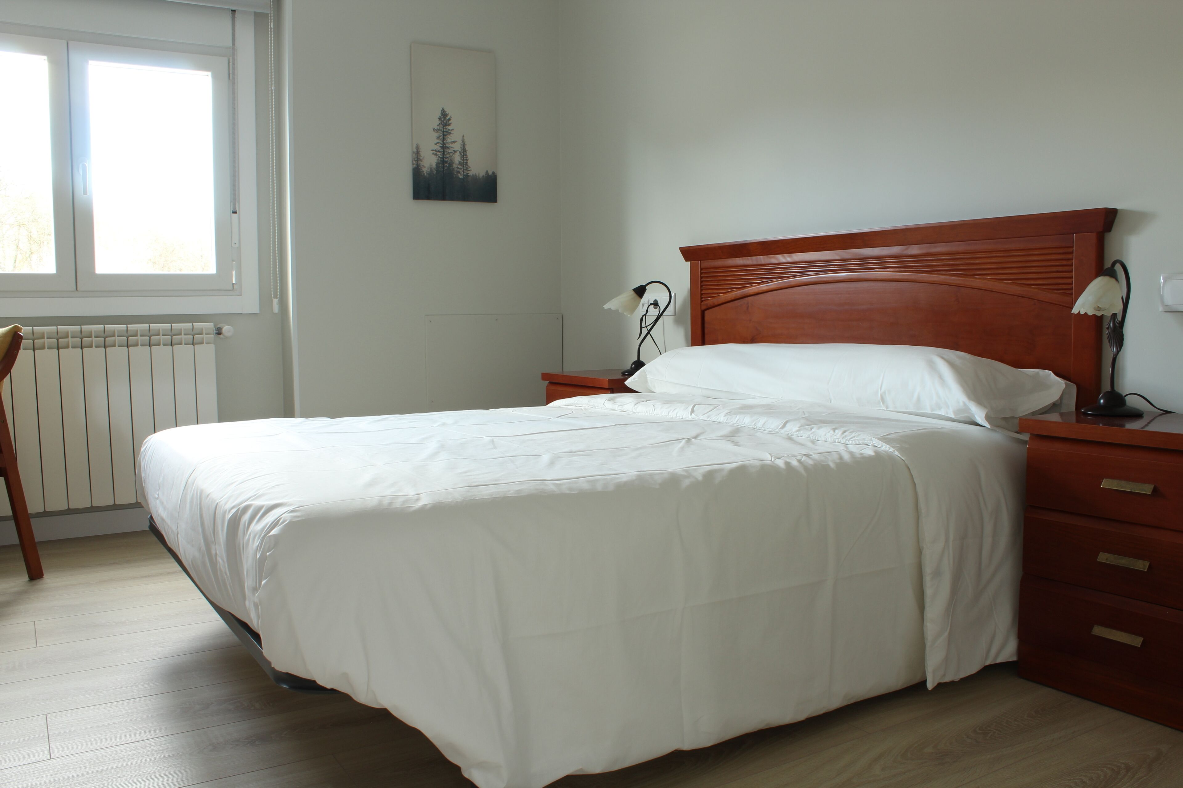 A neatly made bed with white bedding in a room with wooden flooring, a nightstand with a lamp, and a radiator under a window.