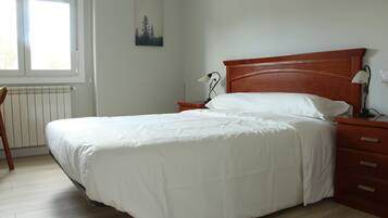 A neatly made bed with white bedding in a room with wooden flooring, a nightstand with a lamp, and a radiator under a window.