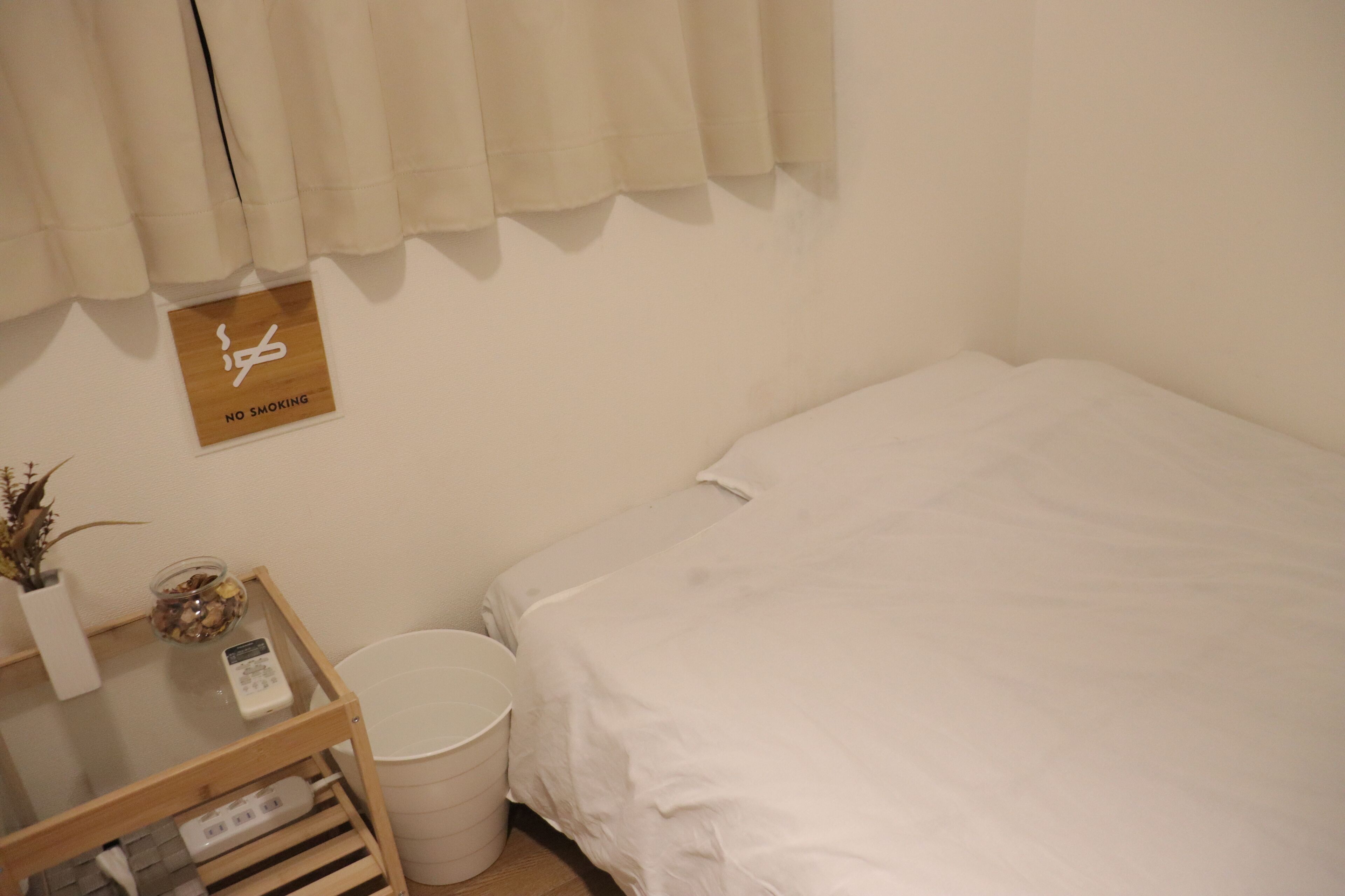A small, clean room with a bed, a bedside table with a lamp, a trash bin, and a 'No Smoking' sign on the wall.
