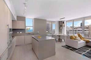 Eine moderne Wohnung mit Küche, Essbereich und Wohnzimmer. Sie verfügt über eine große Kochinsel, einen Esstisch mit Stühlen und ein Sofa mit Blick auf die Stadt.