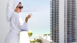 A person in a white robe holding a cup, overlooking a cityscape with a clear view of the ocean and tall buildings. - Wrap Around Balcony|Insane Amenities+ Amazing Pool (Hollywood)