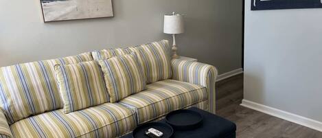 A striped sofa, a small table with a remote control, and a lamp on a white nightstand.