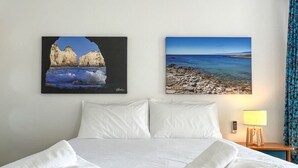 A hotel room with a bed, white linens, two paintings of coastal scenes, a nightstand with a lamp, and a blue curtain.