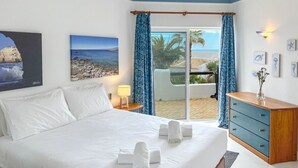 A hotel room with a bed, a dresser, a nightstand, a lamp, and a view of the beach.