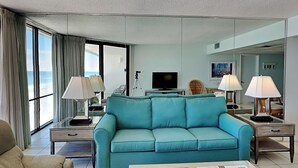A modern hotel room with a teal sofa, a television, and a large window offering a view of the ocean. - Edgewater Windward #603 | Gulf Front + Resort Amenities (Panama City Beach)