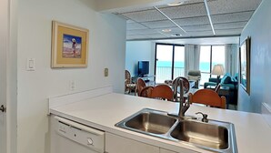 A compact kitchen with a double sink, a dining area with chairs and a table, and a large window offering a view of the ocean. - Edgewater Windward #603 | Gulf Front + Resort Amenities (Panama City Beach)
