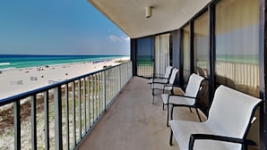 A balcony with a view of the beach, featuring a railing, a few chairs, and a window with curtains.