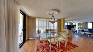 A hotel room with a dining area featuring a glass table and chairs, a large window with curtains, and a view of the ocean.