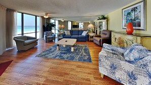 A spacious living room with a large window offering a view of the beach, a blue patterned sofa, a beige armchair, and a wooden coffee table.