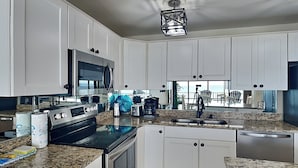 A modern kitchen with white cabinets, granite countertops, stainless steel appliances, and a large window offering a view of the outdoors.