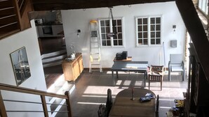 A spacious loft-style living area with a high ceiling, exposed wooden beams, a staircase, a kitchen, and a dining area with a table and chairs.