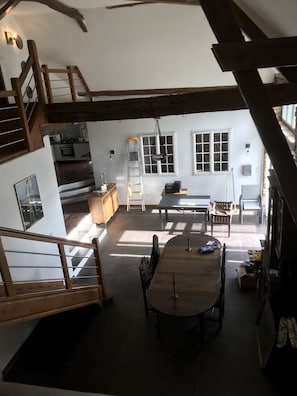 Un vaste espace de vie aménagé dans un style « loft », avec un plafond haut, des poutres apparentes, un escalier, une cuisine et un coin repas comprenant une table et des chaises.