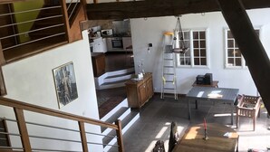 A spacious loft-style room with a high ceiling, exposed wooden beams, a staircase, a dining area with a table and chairs, and a kitchen area with a stove and microwave.