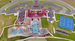 An aerial view of a resort with a central building, multiple swimming pools, tennis courts, and a parking area.