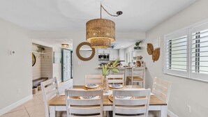 A dining area with a wooden table, white chairs, a woven pendant light, and a kitchen in the background. - 6703 Midnight Pass RD · Siesta Key Condo, Heated Pool, Newly Renovated (Siesta Key)