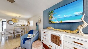 A living room with a large TV, a dining area with white furniture, and a kitchen in the background. - 6703 Midnight Pass RD · Siesta Key Condo, Heated Pool, Newly Renovated (Siesta Key)