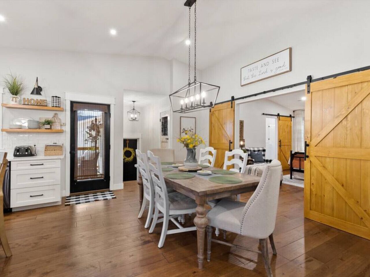 Un comedor con una mesa de madera, sillas blancas y una araña. Hay una zona de cocina con armarios blancos y una puerta negra.