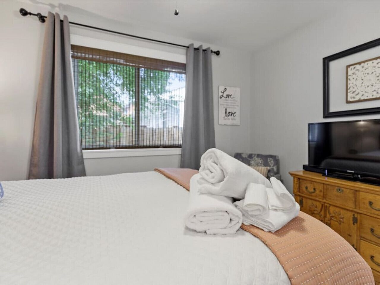 A neatly made bed with a white comforter and a folded towel on the side. There is a television on a wooden dresser and a window with curtains.