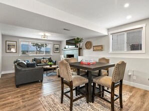 A modern living room with a dark wood floor, a gray sofa, a dining table with four chairs, and a fireplace.