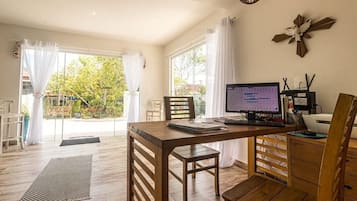 A room with a wooden desk, computer, and chair, with large windows and a view of greenery outside.