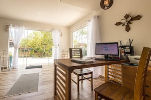 Ein Zimmer mit einem Holztisch, Computer und Stuhl, großen Fenstern und Blick ins Grüne.