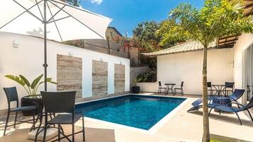 A swimming pool with lounge chairs and a table, surrounded by a tiled patio and a small garden area.