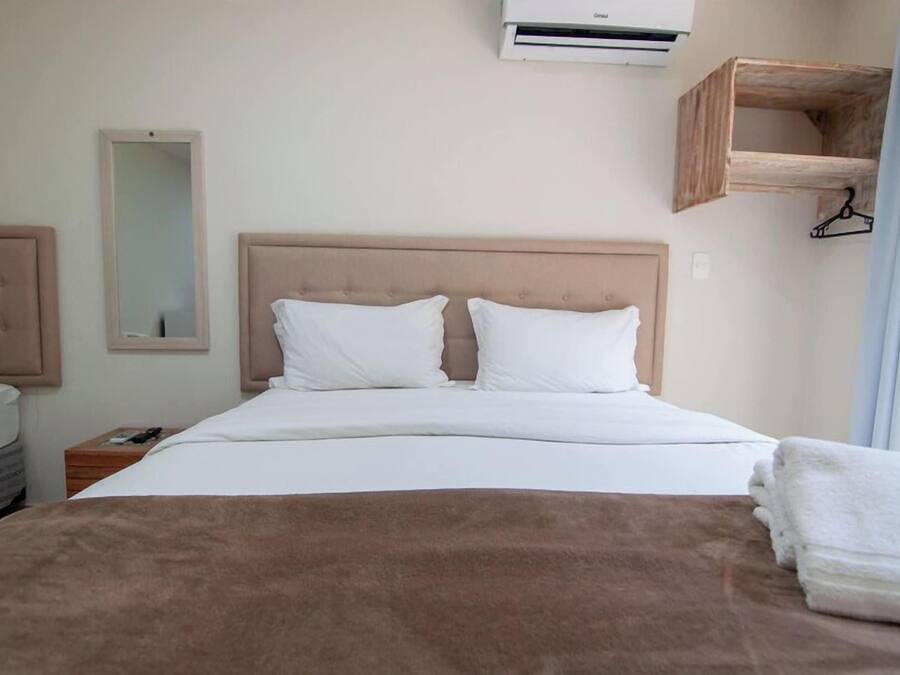 A hotel room with a bed, a bedside table, a mirror, an air conditioning unit, and a wooden shelf.